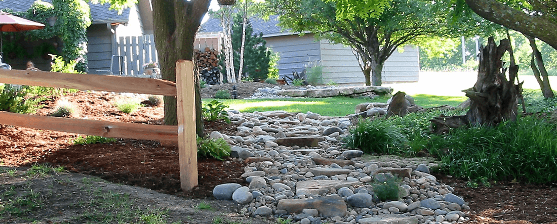 Landscaped yard featuring a wooden fence, decorative stone for drainage, and mulched areas with greenery
