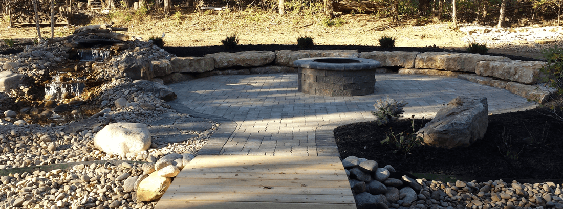 A small, wooden footbridge leading to a stone patio with limestone seating, fire feature in the center, and water feature along the outside.